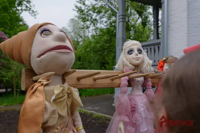 Two large marionette dolls with big eyes and long lashes sit on a light wooden railing outdoors, one in a peach dress and the other in a pink lace dress, with green trees in the background.