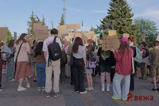 Crowd of people in a public square holding handwritten cardboard protest signs, seen from behind, with trees and street lamps in the background.