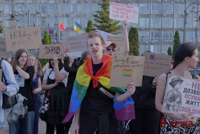 Protesters hold handwritten signs in Ukrainian in a city square; a front-runner wears a rainbow flag sash.