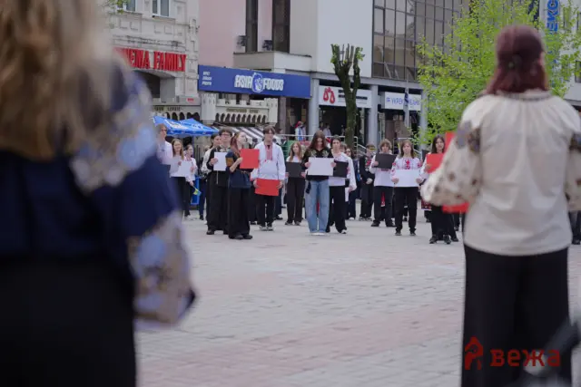 Group of students in traditional clothing holding colored sheets on a city street, with blurred spectators in the foreground.