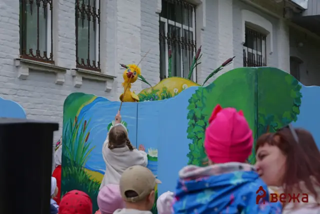 Children in colorful costumes reach toward a yellow bird puppet on a stick above a painted mural at a street parade.