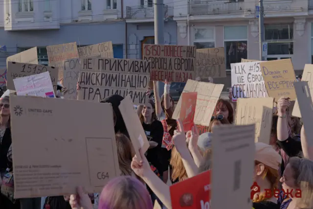 Crowd of protesters in a street, holding large cardboard signs with Cyrillic writing.