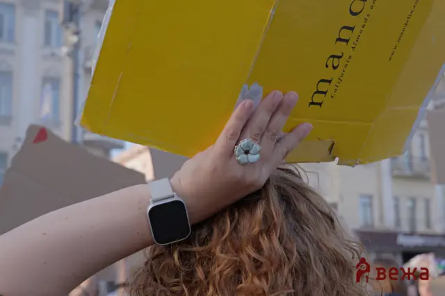 Person with long wavy hair holding a large yellow cardboard box above their head in a city street, wearing a smartwatch and a decorative ring on the hand.