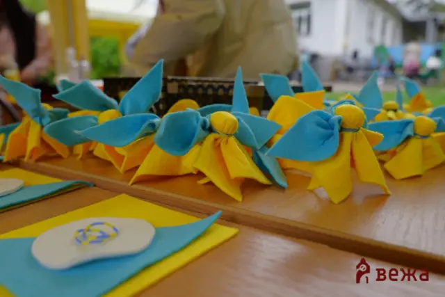 Blue and yellow fabric bows arranged on a wooden table at a craft table, with a blurred busy background and logo in the corner.