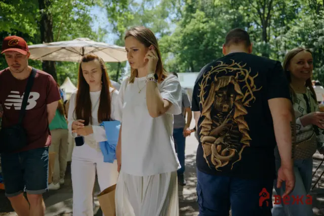 Group of people at an outdoor event in a park; central woman in white speaks on a phone, others around her adapting messages or carrying bags.