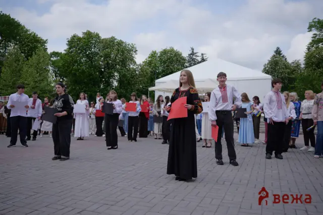 Group of young people in traditional Ukrainian clothing standing on a plaza, holding colored sheets of paper against a backdrop of trees and a white tent.