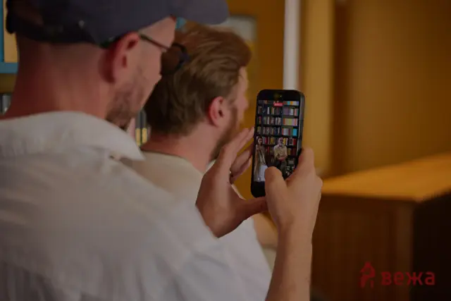Two men filming a scene with a smartphone; the phone screen shows a bookshelf and two people speaking in a room with shelves behind them.