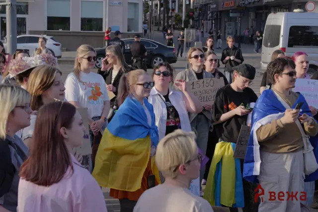 Crowd of protesters on a city street, some wrapped in Ukrainian flags, holding signs; police are visible in the background.