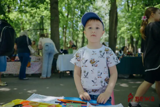 Boy wearing a blue cap at a craft table outdoors, with crayons and paper in a park setting.