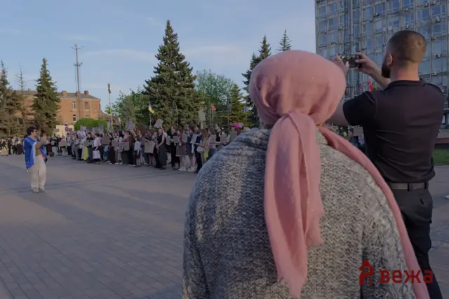 Group of people protesting in a city square, many holding handmade signs, with a photographer and a pink-scarfed person in the foreground.