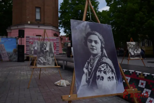 Outdoor art exhibit with large black-and-white portrait on an easel, other portraits visible in the background near a pink building.