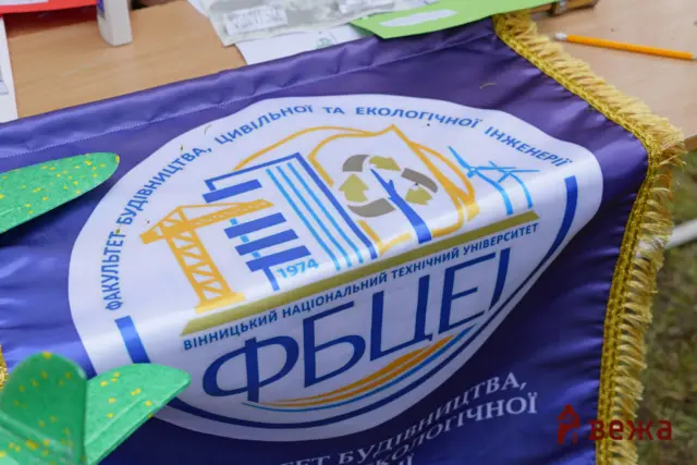 Close-up of a purple banner with a white emblem in blue and yellow and Ukrainian text, on a wooden table; a green decorative leaf and a pencil nearby.