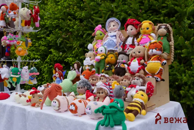 Assorted handmade crocheted stuffed animals and dolls displayed on a white table at an outdoor market, with greenery in the background.
