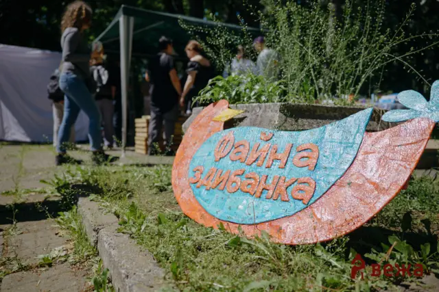 Colorful, weathered sign with Cyrillic text in the foreground; blurred people chatting near a tent in the background, sunny outdoor scene.