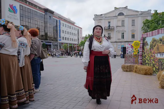People in traditional outfits with embroidered skirts and headscarves stand in a city square during a cultural festival; a woman in the center wears multiple bead necklaces.
