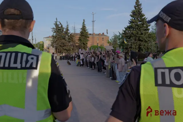 Protesters holding placards march along a city street, with two police officers in high-visibility vests in the foreground and a Ukrainian flag in the background.