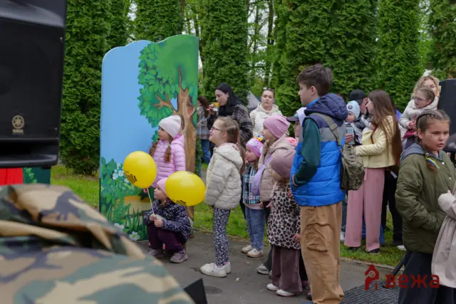 Group of children and adults watching an outdoor event beside a large blue tree-cutout backdrop; some kids hold yellow balloons and wear colorful jackets/hats.