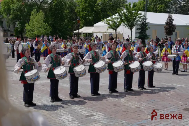 Group of young drummers in Ukrainian folk costumes performing in a city square during a parade, with onlookers and tents in the background.
