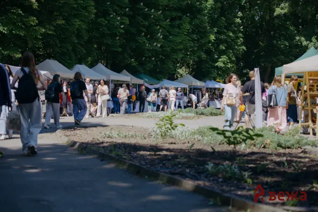 People browse stalls at an outdoor market with white tents and lush trees in the background.