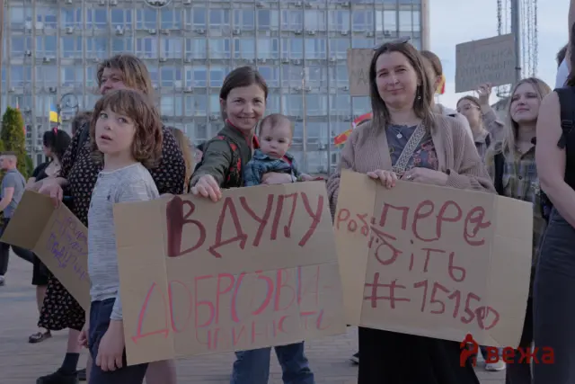 Group of protesters in an urban square holding large cardboard signs with red Cyrillic text; a building with many windows in the background.