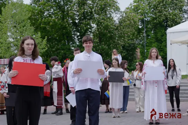Group of young people in traditional Ukrainian embroidered outfits holding signs at an outdoor ceremony. The foreground shows three individuals with large white and red placards, with a crowd and green trees in the background.