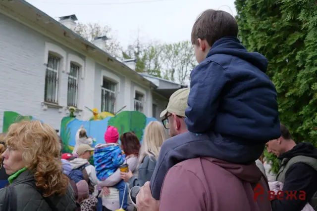 Crowd at an outdoor festival; a man in a blue hoodie carries a child on his shoulders, with a colorful mural and white brick building to the left.