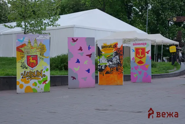 Outdoor festival scene with white tents and colorful display boards featuring city illustrations; a person on a bicycle with a yellow Glovo backpack appears on the right.