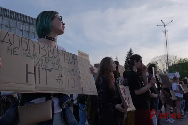 Protesters hold large handwritten cardboard signs at an outdoor rally, faces turned to the side, city street and streetlights in the background.