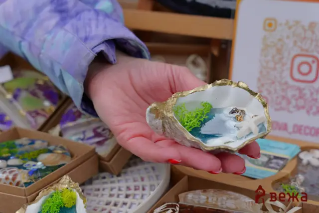 Hand holding a decorated seashell filled with blue paint and bright green moss, shown in a craft display.