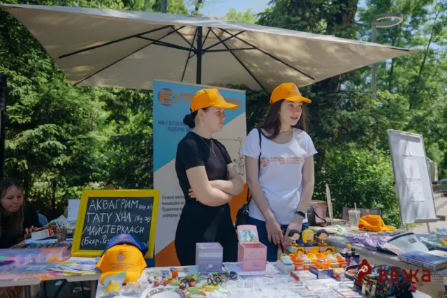 Two women in orange caps run an outdoor craft stall under a large umbrella, with handmade items and a chalkboard sign on the table in front of them.