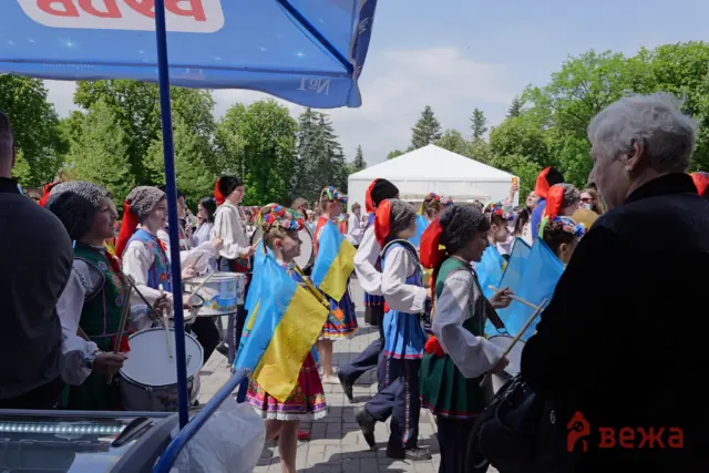 Group of children in Ukrainian folk costumes marching with drums and blue-yellow flags during an outdoor festival.