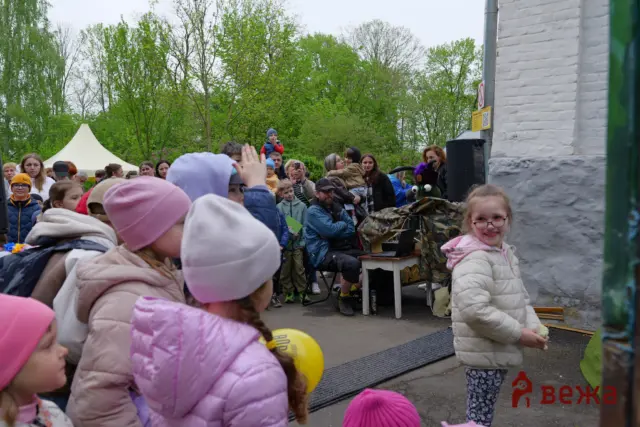 Crowd of children at an outdoor event, wearing colorful jackets, with trees and a white tent in the background.