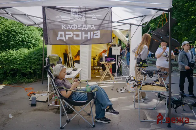 Outdoor drone exhibition booth with a gray banner reading 'КАФЕДРА ДРОНІВ' and people handling equipment; trees in the background.