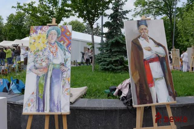 Outdoor art exhibit with tall painted portraits on wooden easels; a colorful portrait of a woman in a headscarf on the left and a man in period clothing on the right, with tents and people in the background.