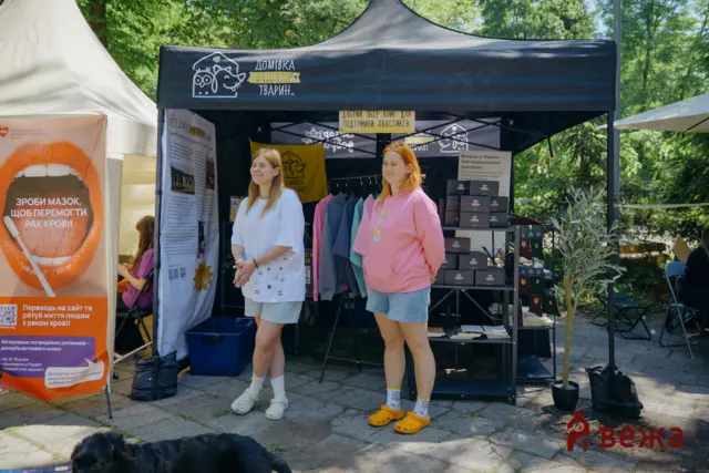 Two women stand at an outdoor charity booth under a black canopy, with banners and merchandise visible behind them; a black dog rests on the ground in front.
