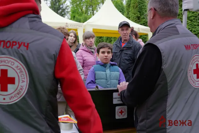 Boy in a blue-green puffer jacket at a humanitarian aid booth, flanked by adults in Grey Red Cross vests near white tents outdoors.