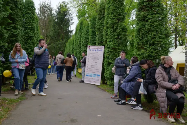 People gathered along a park path with a tall evergreen hedge and a large education poster nearby.