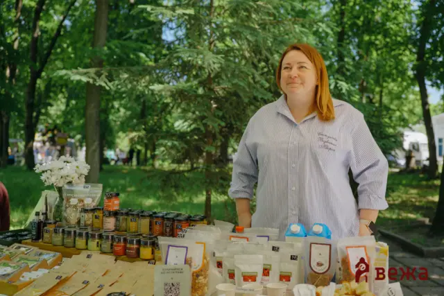 Woman standing at an outdoor market table with packaged foods and jars, greenery in the background.