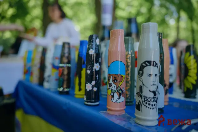 Row of colorful painted bottles with dog illustrations on a blue table outdoors, with green trees in the background.