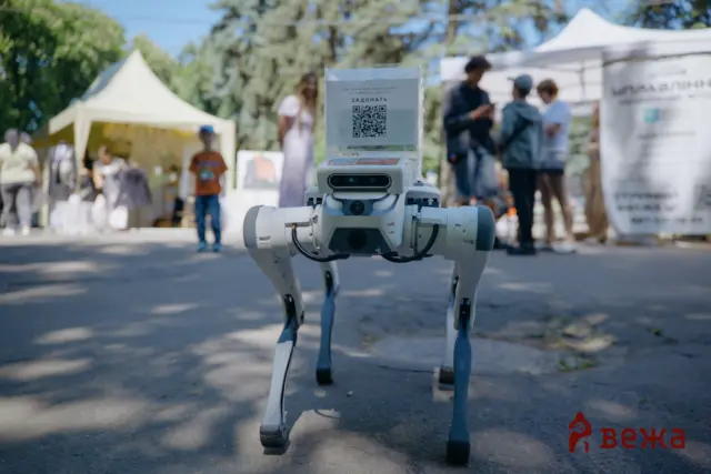 Humanoid robot standing outdoors with a QR code sign on its head; blurred event tents and people in the background behind it.