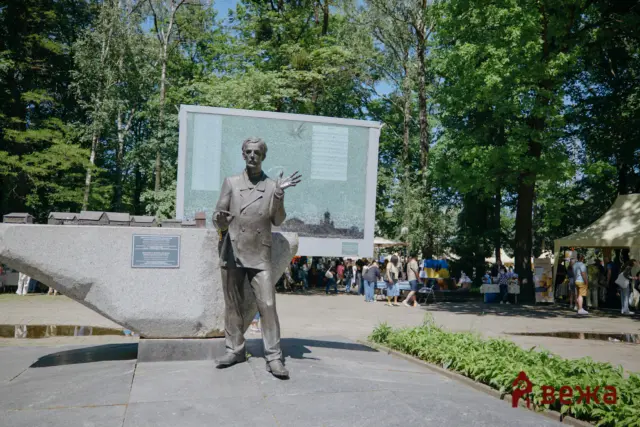 Bronze statue of a man in a suit gesturing with his right hand in a public square, with a large glass-information panel behind and trees in the background.