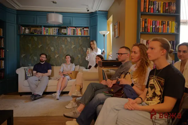 Group of people sit in a blue-walled living room; a woman stands by a yellow wall using a smartphone to record or take a photo.