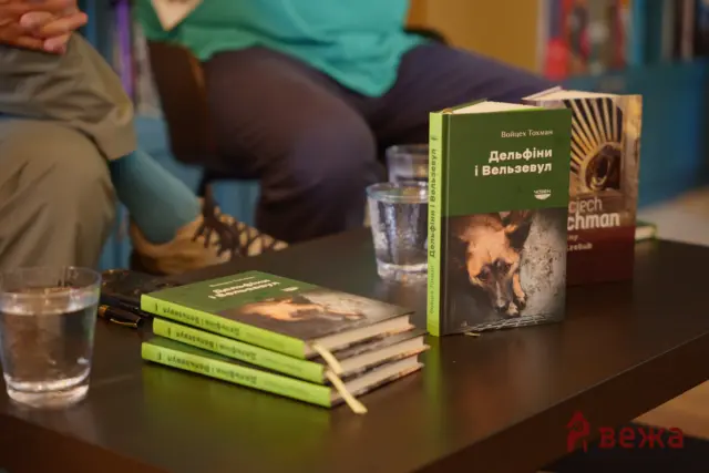 Green paperback books with Cyrillic titles on a dark table, a blurred person in a teal shirt in the background.