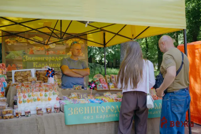Vendor under a yellow canopy selling colorful candies and baked goods at an outdoor market; two customers browse the stall.