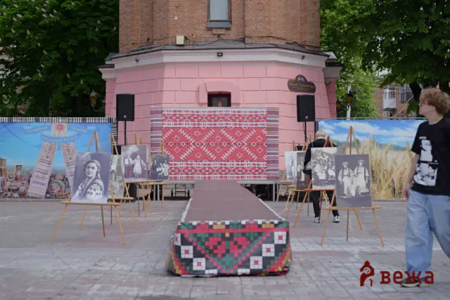 Outdoor cultural exhibit with framed vintage photos on easels, displayed in front of a pink brick building and colorful woven textiles hanging on a wall behind.