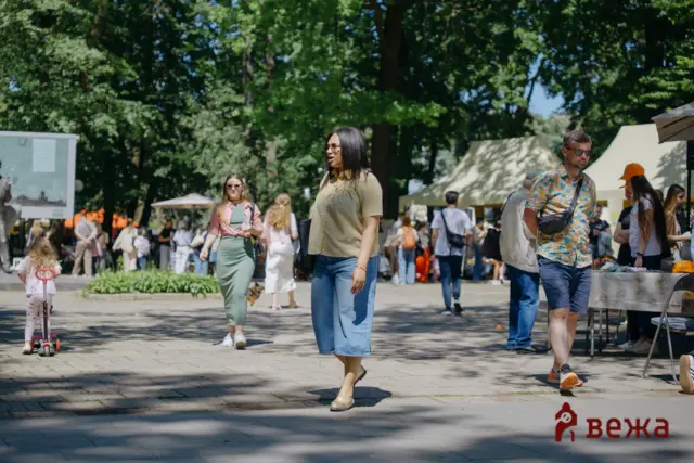 People strolling in a sunlit park with tents and trees in the background; foreground woman in a beige checkered shirt walks forward wearing sunglasses.