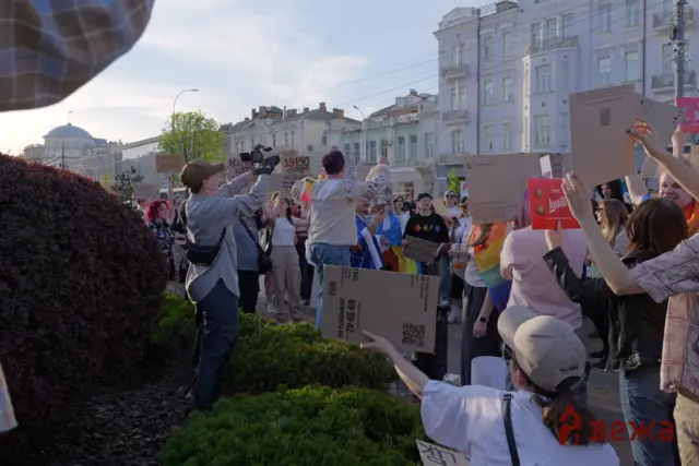 Crowd of protesters on a city street holding cardboard signs, some with QR codes and rainbow flags visible against pastel buildings.