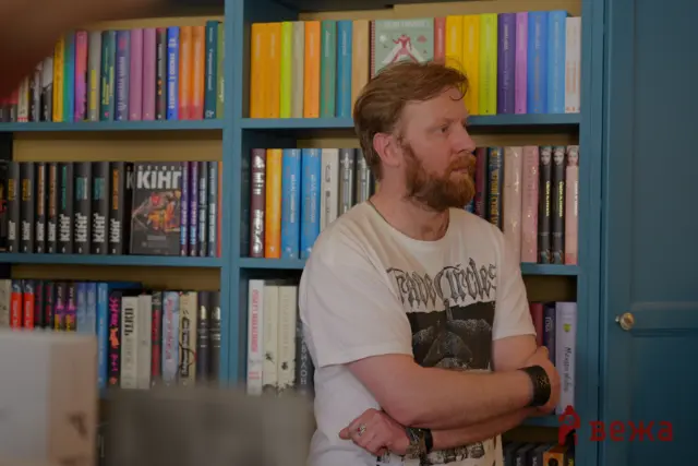 Bearded man in a white graphic tee stands in front of blue bookshelves filled with colorful books, looking to his left.