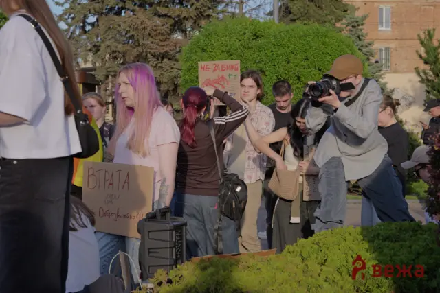 Group of young protesters holding cardboard signs in a park, with a photographer aiming a camera nearby.