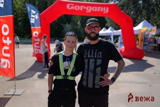 Two men posing together at an outdoor event under a large red inflatable arch; one wears a neon safety vest and race bib, the other in a black shirt and cap.